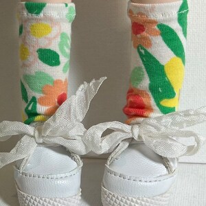 May include: White sneakers with white laces tied in a bow. The sneakers have a white sole with a textured pattern. The person is wearing white socks with a floral pattern in shades of green, yellow, orange, and pink.