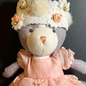 May include: A gray plush toy wearing a white lace hat with pink and white flowers. The toy is wearing a pink dress with ruffles.