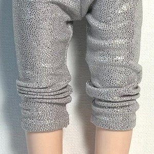 May include: Silver glitter leggings for a doll. The leggings are a light gray color with silver glitter woven into the fabric. The leggings are cuffed at the bottom.