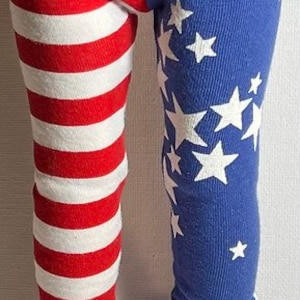 May include: Red, white, and blue leggings with a striped pattern on one leg and a star pattern on the other leg. The leggings are worn by a doll with white sneakers.