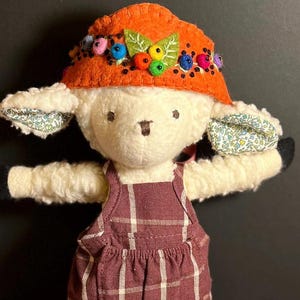 May include: A white plush lamb doll wearing a brown and orange felt hat with colorful beads and a brown and white plaid overall dress.