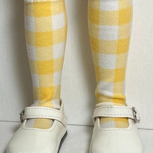 May include: A pair of white patent leather shoes with a buckle closure. The shoes are worn with yellow and white checkered knee-high socks.