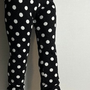 May include: Black and white polka dot leggings for a doll. The leggings are made of a soft, stretchy fabric and have a comfortable fit. The doll is wearing white sneakers with black accents.