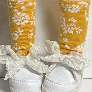 May include: A pair of white sneakers with white lace-up bows and yellow socks with a white floral pattern.