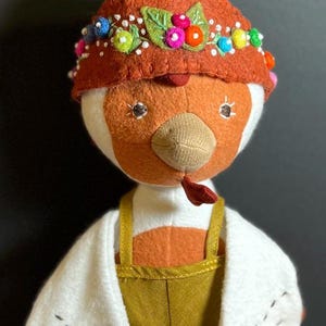 May include: A plush toy chicken wearing a brown felt hat with colorful beads and leaves. The chicken is wearing a white cardigan and brown overalls.