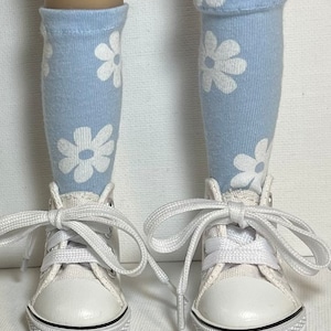 May include: A pair of white high-top sneakers with white laces. The sneakers are worn by a doll with light blue socks with white daisy patterns.
