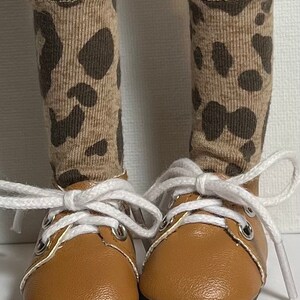 May include: A pair of brown leather lace-up shoes with white laces. The shoes are worn by a doll with brown and tan leopard print socks.