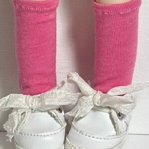 May include: A pair of white sneakers with pink socks and white ribbon laces. The sneakers have a white rubber sole.