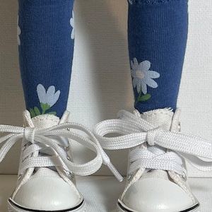 May include: A pair of white canvas high-top sneakers with white laces. The sneakers are worn by a doll with blue socks with white flower patterns.