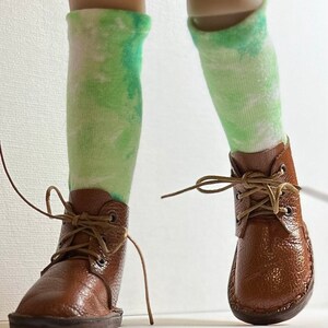 May include: A pair of brown leather ankle boots with laces tied. The boots are worn by a doll with green and white tie-dye socks.
