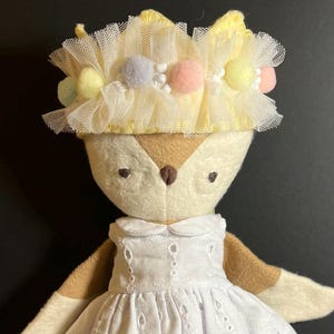 May include: A plush toy fox wearing a white dress with eyelet lace and a yellow tulle crown with pastel pom poms.