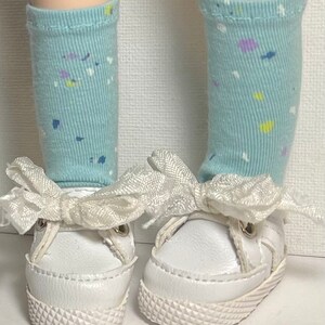 May include: A pair of white canvas shoes with white laces and a white bow on each shoe. The shoes are worn by a doll with light blue socks with a white, yellow, and purple confetti pattern.