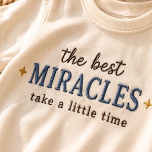 May include: A cream-coloured short-sleeve t-shirt featuring dark brown and blue embroidered text that reads the best MIRACLES take a little time, accented by two small gold-coloured embroidered stars on either side of the word MIRACLES.