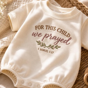 Op de afbeelding: Een crèmekleurige baby-romper met korte mouwen en geborduurde tekst FOR THIS CHILD we prayed en 1 SAMUEL 1:27, geaccentueerd met een klein groen bladontwerp. Het kledingstuk heeft een geribbelde ronde hals en drukknopen aan de onderzijde.