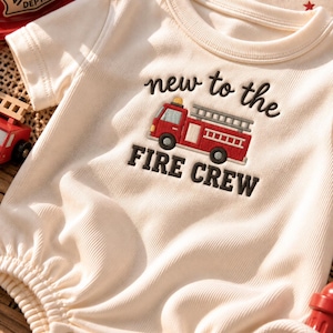 May include: A cream-colored ribbed short-sleeve bodysuit featuring an embroidered red fire truck and the text "new to the FIRE CREW" in dark brown lettering. The garment has a round neckline and a snap-button closure at the bottom hem.