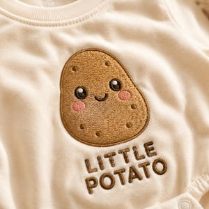 Peut inclure: Un body à manches longues de couleur crème orné d'une pomme de terre brodée en marron avec un visage souriant, des joues roses et des yeux sombres, situé au-dessus du texte brodé en marron indiquant LITTLE POTATO dans une police sans empattement.