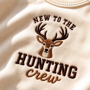 Könnte beinhalten: Ein cremefarbenes geripptes Shirt mit einem gestickten braunen Hirschkopf in der Mitte, umgeben von dem Text NEW TO THE darüber und HUNTING crew darunter in dunkelbrauner Block- und Schreibschrift. Das Motiv ist direkt auf den Stoff gestickt.