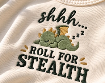 Puede incluir: Una prenda de tela acanalada color crema con un bordado de un dragón verde durmiendo con alas doradas. El texto dice shhhh... ROLL FOR STEALTH en hilo verde oscuro y dorado, acompañado de pequeños símbolos zzz y detalles en color oro.