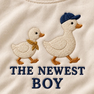 Peut inclure: Un vêtement en tissu blanc cassé avec une broderie représentant deux canards couleur crème. L'un porte un foulard jaune autour du cou, l'autre une casquette bleu marine. Sous les canards, le texte THE NEWEST BOY est brodé en bleu marine.