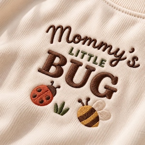 Könnte beinhalten: Ein cremefarbenes, geripptes Sweatshirt mit dunkelbraunem Sticktext, der Mommy's LITTLE BUG lautet. Unter dem Text befinden sich ein gestickter roter Marienkäfer, ein kleiner grüner Grashalm und eine gelb-braun gestreifte Biene mit weißen Flügeln.