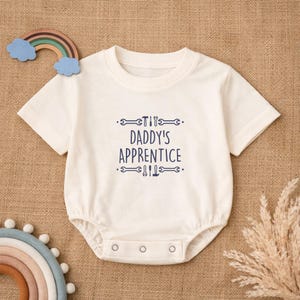 Puede incluir: Body de bebé color crema con mangas cortas y cierre de corchetes. El texto "DADDY'S APPRENTICE" está impreso en azul marino, junto con ilustraciones de herramientas. Un arcoíris de madera y una ramita de hierba seca están en el fondo.