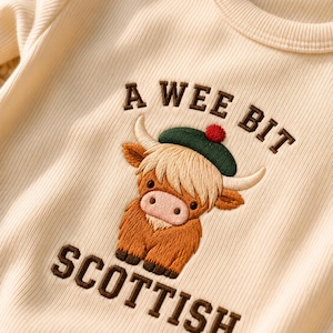 Pode incluir: Uma camisola de manga comprida canelada de cor creme com um bordado de uma vaca das Terras Altas castanha que usa uma boina verde com um pompom vermelho. O texto "A WEE BIT" está bordado acima e "SCOTTISH" está bordado abaixo.