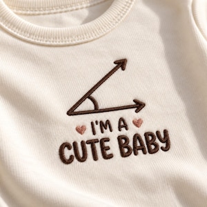 May include: An off-white baby bodysuit featuring dark brown embroidery of an acute angle symbol above the text "I'M A CUTE BABY" in dark brown, flanked by two small light pink embroidered hearts. The garment has a ribbed crew neckline.