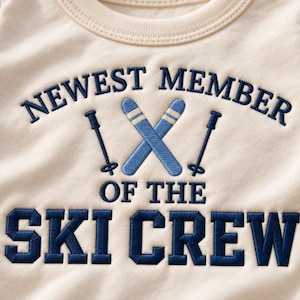 Op de afbeelding: Een crèmekleurig shirt met donkerblauw borduurwerk met de tekst NEWEST MEMBER OF THE SKI CREW. Het ontwerp bevat een geborduurde afbeelding van twee gekruiste blauwe ski's en twee skistokken tussen de tekstregels door.
