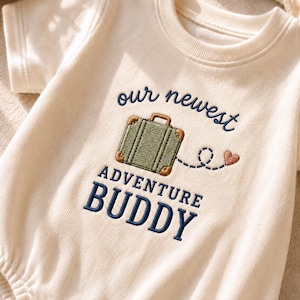 May include: A cream-coloured short-sleeve bodysuit featuring embroidered text that reads our newest ADVENTURE BUDDY. The design includes a green suitcase with brown corners and a dashed line leading to a small pink heart, all in dark blue thread.