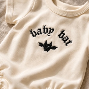 以下が含まれることがあります： クリーム色の半袖ベビーボディスーツ。ゴシックフォントでbaby batと刺繍された黒いテキストと、その下に小さな黒いコウモリの刺繍が施されています。リブ編みのクルーネックとスナップボタンが付いています。