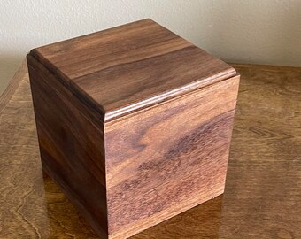 May include: A square wooden box with a dark brown finish. The box has a smooth surface and is made of solid wood.