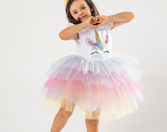 May include: A young girl wearing a white unicorn dress with a rainbow tulle skirt and a unicorn headband. She is making a heart shape with her hands.