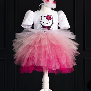 Puede incluir: Un tutú de tul blanco con un efecto degradado rosa. El tutú está en capas y tiene una capa superior blanca con un gráfico de Hello Kitty rosa. El tutú está en un soporte blanco sobre un fondo negro.