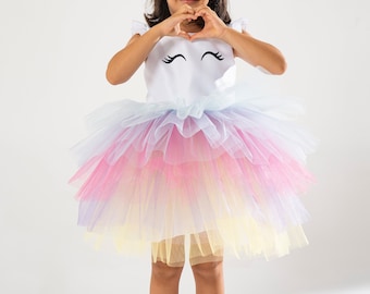 May include: A child's unicorn costume featuring a white top with a unicorn face design and a multi-colored tulle skirt. The costume includes a gold horn headband with flowers and white shoes.