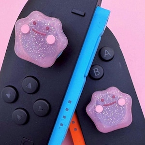 以下が含まれることがあります： 黒いゲームコントローラーのクローズアップ。青いサイドレールと、ピンク色のキラキラしたDitto型のサムグリップが2つあります。サムグリップには頬のピンク色の円と小さな笑顔があります。コントローラーにはAとBとラベルされた黒いボタンがあります。