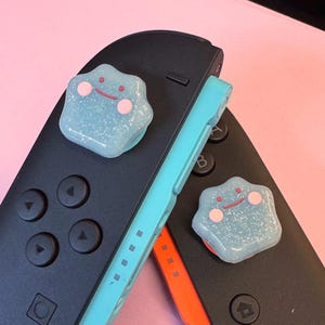 以下が含まれることがあります： 黒いゲームコントローラー2つ、水色のケース、Ditto型のサムグリップ2つ。 グリップは水色で、ピンクの頬と赤い笑顔があります。 コントローラーはピンク色の背景にあります。