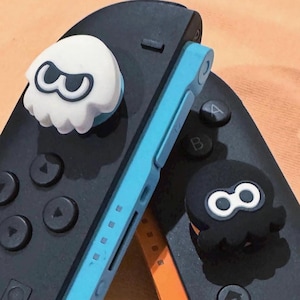 May include: Black and blue game controller with white and black character-shaped thumb grips. The controller has a blue frame and black buttons. One thumb grip is white with a black design, and the other is black with a white design.