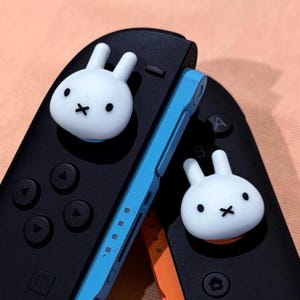 以下が含まれることがあります： 青いアクセントが付いた2つの黒いゲームコントローラー。それぞれに白いウサギ型のサムグリップが付いています。ウサギは黒い点目の目と口の黒い十字架があります。 コントローラーは、桃色の背景に配置されています。