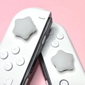 Puede incluir: Primer plano de dos mandos de juego blancos con agarres para el pulgar en forma de estrella. Los mandos tienen detalles en negro y botones blancos. El fondo es de color rosa liso.