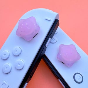 以下が含まれることがあります： ピンクの星型のボタンカバーが付いた白いニンテンドースイッチコントローラー2つ。カバーはキラキラした仕上げになっています。