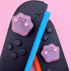 Op de afbeelding: Twee roze, glinsterende Ditto-vormige accessoires liggen op een zwarte gamecontroller. Er is ook een blauwe controllergrip aanwezig. De accessoires hebben roze wangen en een lachend gezicht. De achtergrond is effen roze.
