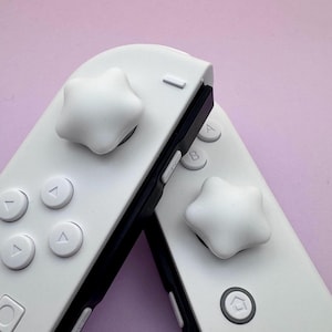 May include: Two white game controllers with star-shaped thumbstick replacements. The controllers have black accents and various button designs, including directional arrows and lettered buttons. The background is a solid light purple.