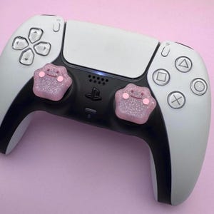 May include: A white and black PlayStation 5 controller with pink glitter paw print-shaped buttons.