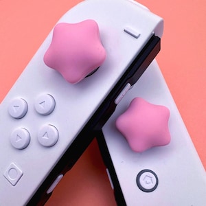 Puede incluir: Primer plano de dos mandos de juego blancos con agarres para el pulgar en forma de estrella rosa. Los mandos tienen detalles en negro y varios botones blancos. El fondo es de color melocotón liso.