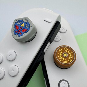 Puede incluir: Dos mandos Nintendo Switch Joy-Con con tapas de botones personalizadas. La tapa izquierda presenta un escudo gris y azul con un diseño rojo y amarillo. La tapa derecha presenta un diseño circular marrón y dorado.