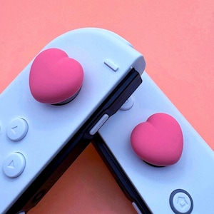 Puede incluir: Dos mandos Joy-Con blancos para Nintendo Switch con tapas de botones en forma de corazón rosa.