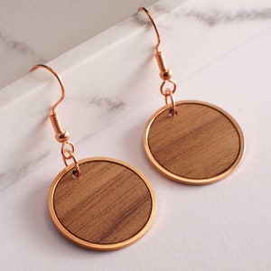 Può includere: Un paio di orecchini rotondi in legno con finitura oro rosa. Gli orecchini sono in legno con un motivo a venature naturali e hanno un cerchio in metallo oro rosa.
