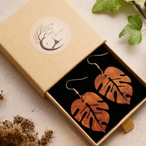 Könnte beinhalten: Ein Paar Holzohrringe in Form von Monstera-Blättern. Die Ohrringe sind braun und haben eine natürliche Holzmaserung. Sie hängen an goldfarbenen Haken in einer kleinen schwarzen Samtbox.