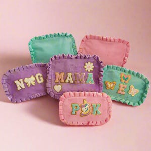 May include: A collection of five rectangular pouches with ruffled edges in pastel shades of pink, purple, and teal. The pouches feature decorative patches including the text MAMA, the letters N, G, P, K, E, a bow, a heart, a flower, a unicorn, and butterflies.