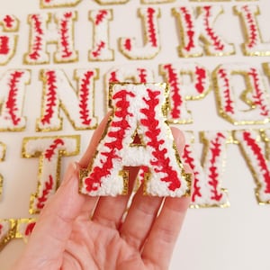 May include: A white felt letter A with red stitching, outlined in gold, shaped like a baseball. The letter is part of a set of alphabet patches.